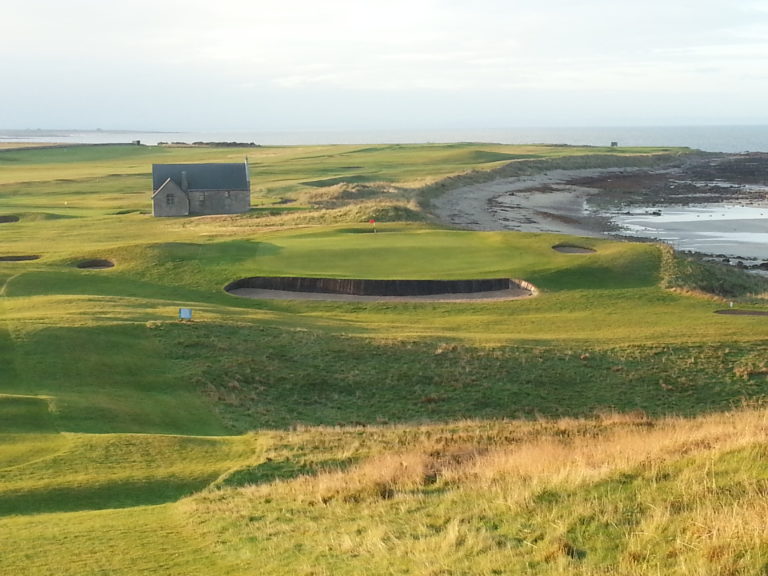 The view down a fairway overlooks a green, The North Sea and an old building, Crail Golfing Society, Fife, Scotland, United Kingdom