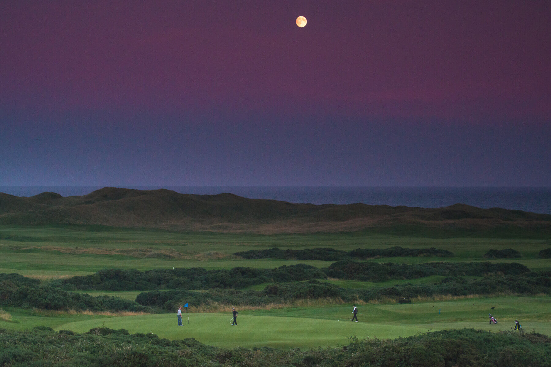 Twilight image of golfers on the 18th green under a twilight sky and pale moon at Cruden Bay Golf Club, Aberdeenshire, Scotland