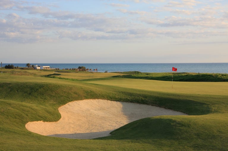 Image depicting a bunker next to a green with a red flag and ocean in the background, Verdura Resort, Sicily, Italy