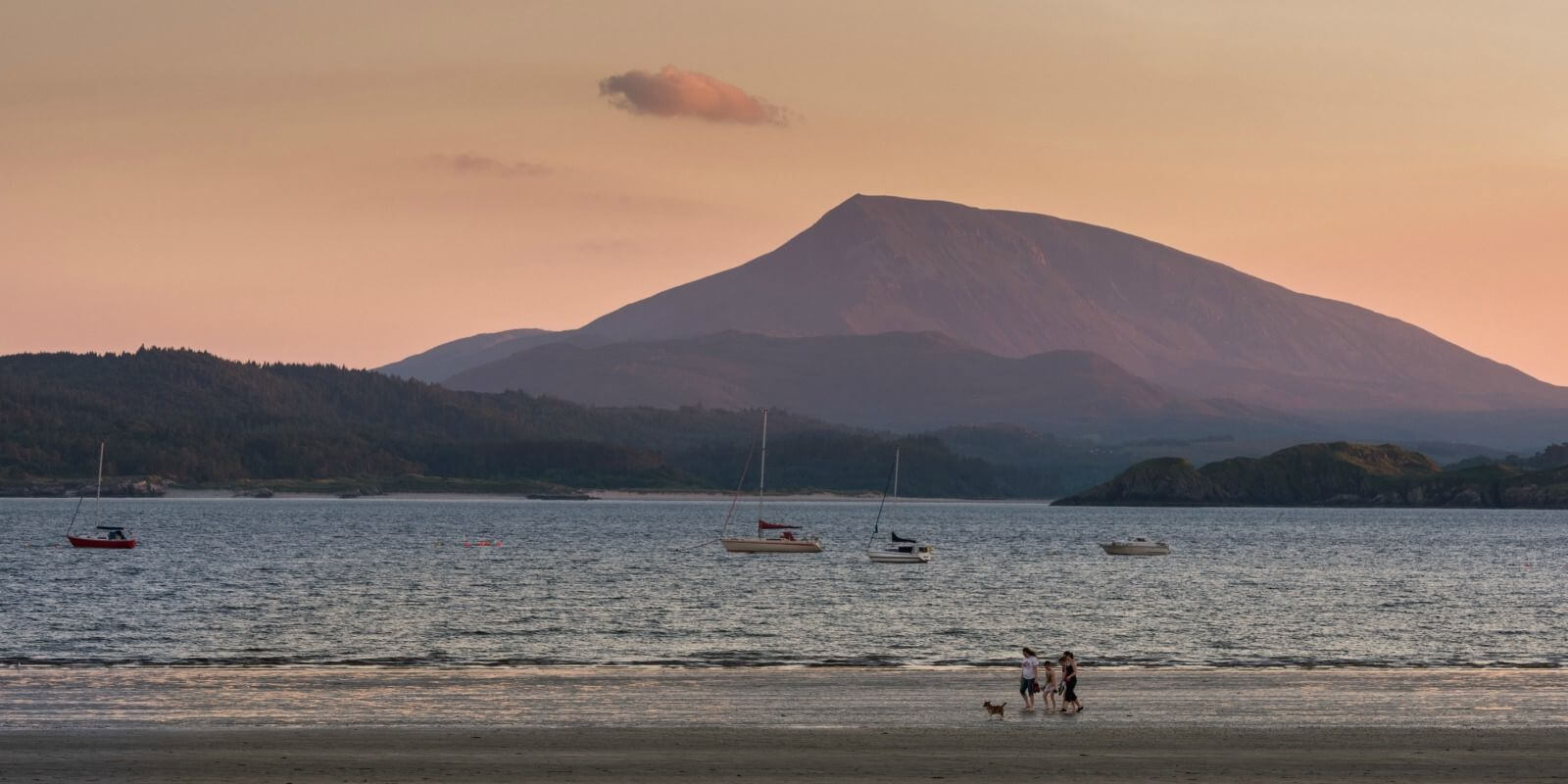 A family walks a dog at the beach with a mountainous background