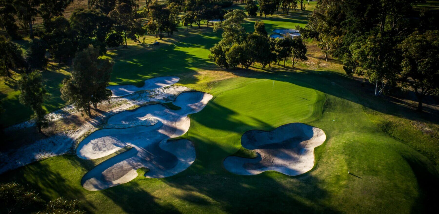 Large native trees create dark shadows on sandy bunkers and a green at Commonwealth Golf Club, Melbourne