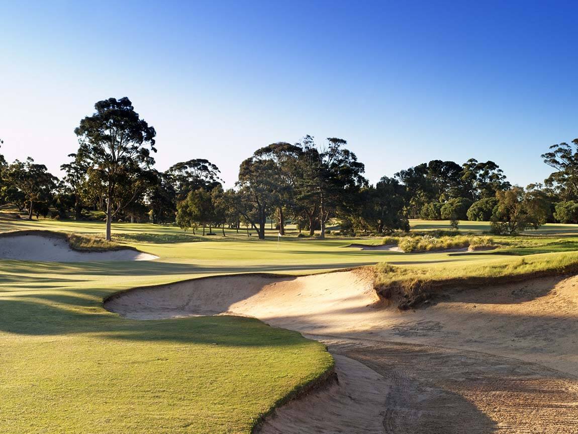 View of a fairway leading down to a green with large trees under a blue sky at The Metropolitan Golf Club, Melbourne